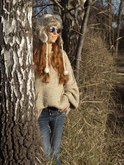 Slender pretty woman with fur hat and sweater, red hair and warm winter colors between birch forest