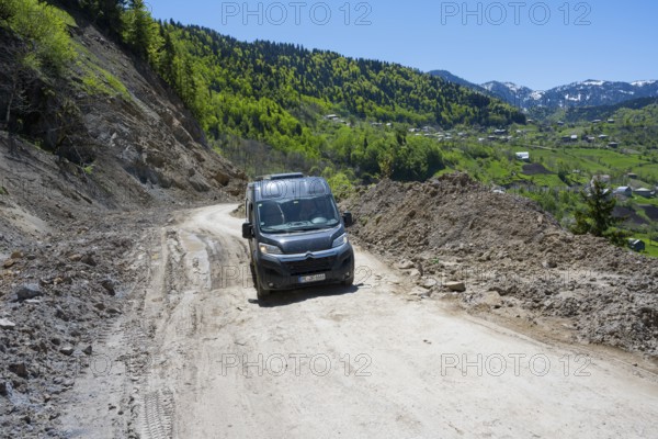 A van drives on a winding mountain road surrounded by wooded hills under blue skies, campers on the