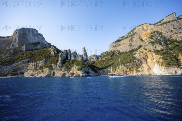 Excursion boat off picturesque rocky coast, cliffs with caves in morning light, L'Aguglia rock pin,