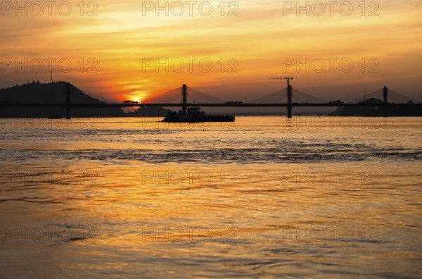A boat sails on the river Brahmaputra during sunset, with the silhouette of a cable-stayed bridge