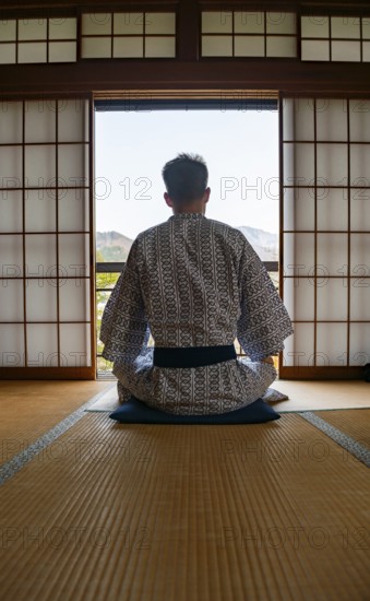 Young man wearing kimono sitting in traditional Japanese living room with tatami mats and shoji