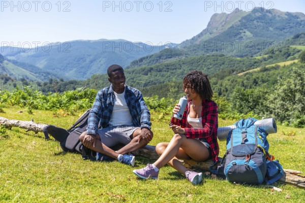 Two hikers are taking a break during their mountain trek, sitting on a log, drinking water and