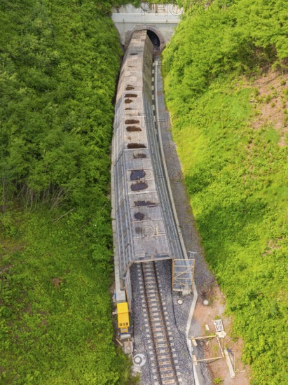 Aerial view of a railway tunnel stretching through green hills and a dense forest, construction of