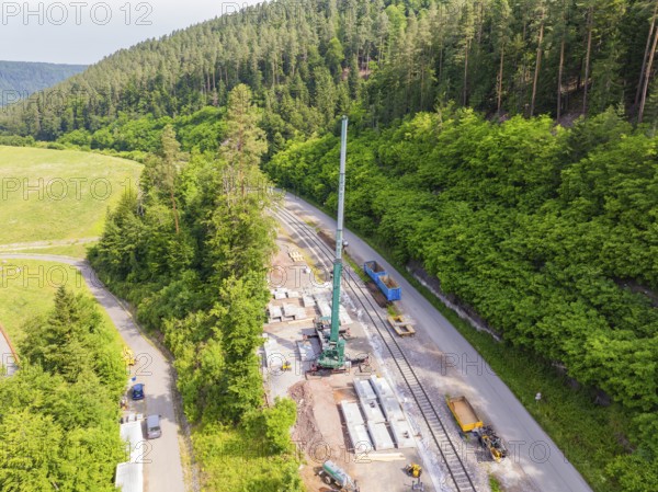 Construction work along a railway line in a green forest area, crane and concrete blocks visible,