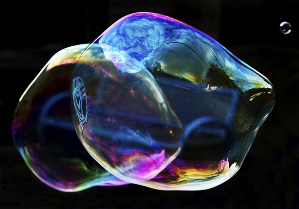 Irregular, iridescent soap bubble in the colours of the rainbow against a black background,