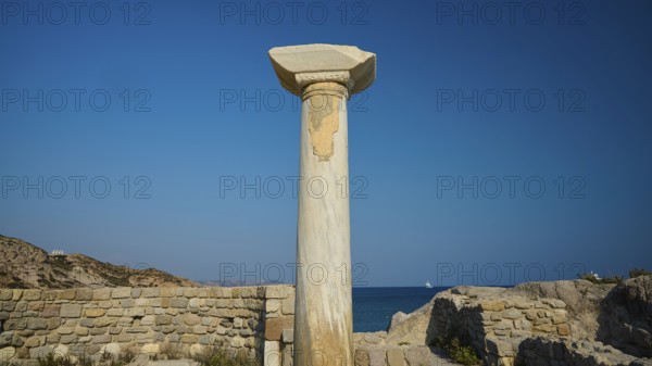 Freestanding stone column in front of a historical site with blue sky in the background, Agios