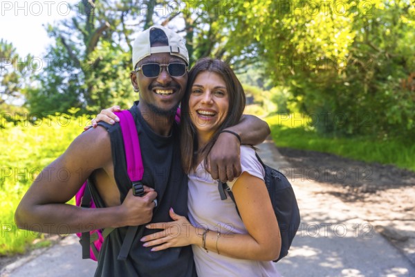 Interracial couple wearing backpacks and embracing while enjoying a hike on a sunny nature trail