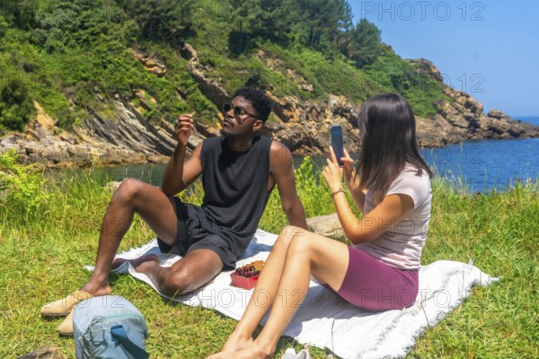 Young woman takes a photo of her friend during a scenic picnic by the sea, capturing memories of