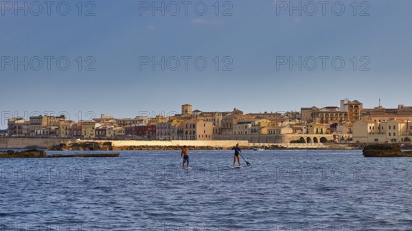 Two paddleboarders on the sea in front of a town with historical buildings under a blue sky, boat