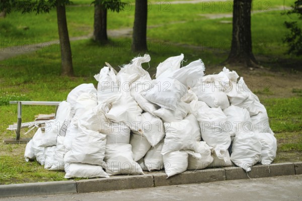 Multiple white heavy-duty garbage bags filled with waste material are piled up in a green park area