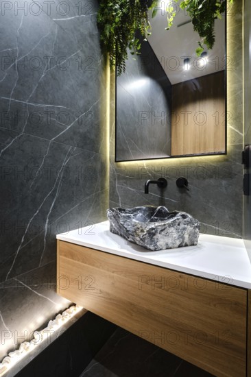 A sleek bathroom with a striking stone sink mounted on a wooden cabinet. The walls are tiled with