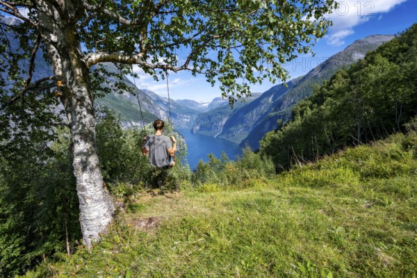 Young man swinging on a tree, view of idyllic fjord landscape of the Geirangerfjord at Blomberg