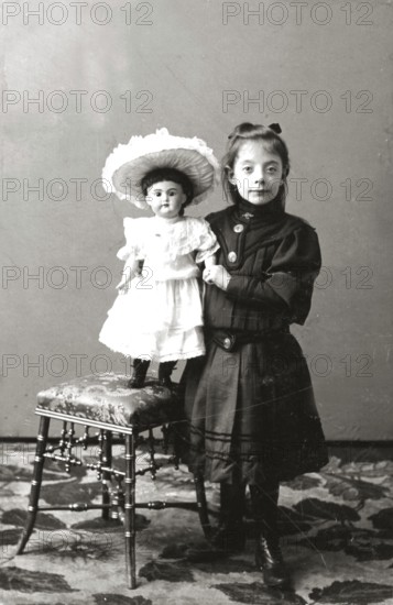Girl doll dresses, 1930s, girl standing next to a chair carrying a doll in a dress, both in