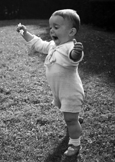 Little boy learning to walk, 1930s, Toddler stands happily in the meadow and raises his right arm,