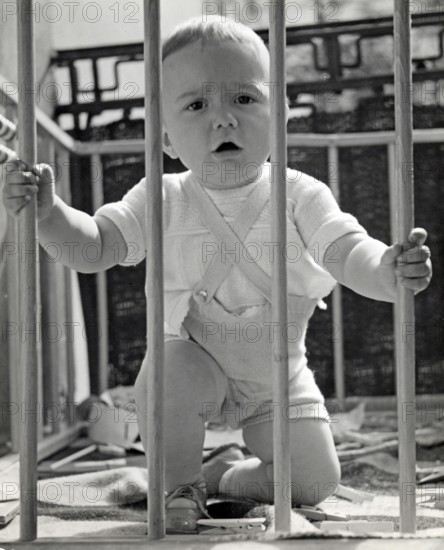 Little boy in a playpen, 1930s, Toddler kneeling in a cot and holding on to the wooden railing,