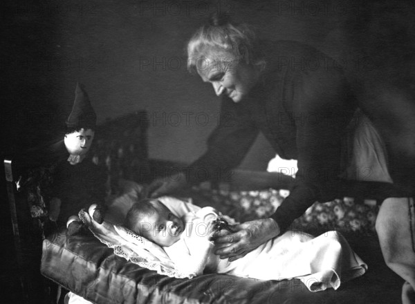 Grandma with grandson, 1930s, An elderly woman lovingly cares for a baby in a cradle, Historical