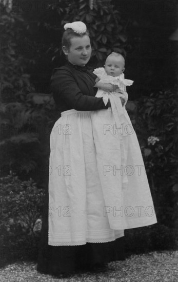 Nanny holding a baby in her arms, ca. 1910, Woman in Victorian dress holding a baby in the garden,