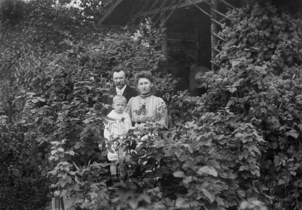 Family standing at a wooden hut, 1920s, adults with child standing in front of a house surrounded
