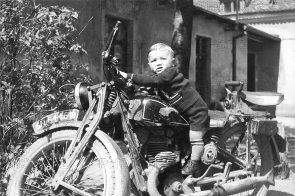 Child on a motorbike, 1930s, Small child sitting smiling on an old motorbike in a rustic setting,