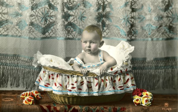 Baby sitting in basket, 1905, A baby sits in a decorative basket draped with colourful fabric,