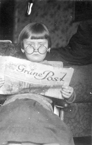Girl's newspaper, 1920s, A child with glasses reads a newspaper with interest, sitting comfortably