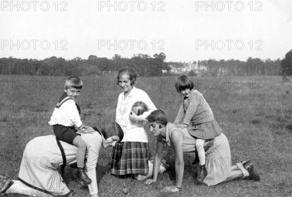 Two children riding on the backs of their mothers, 1930s, Children playing in the meadow with two