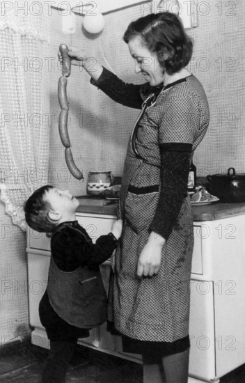 Mother feeding sausages to her son, 1930s, A woman shows sausages to a happy child in a cosy