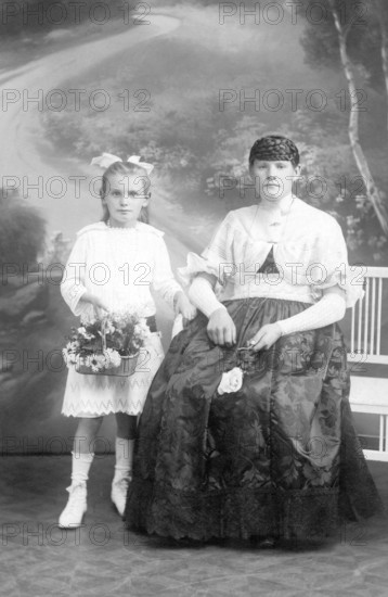 Mother and child, 1930s, A girl in a white dress holding flowers, next to a seated woman in a