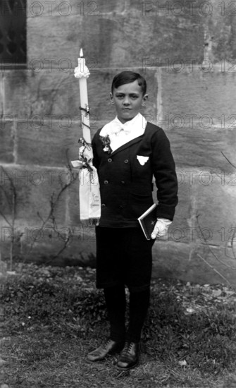 Boy has communion, 1920s, A boy stands with a communion candle and a book in front of a stone wall,