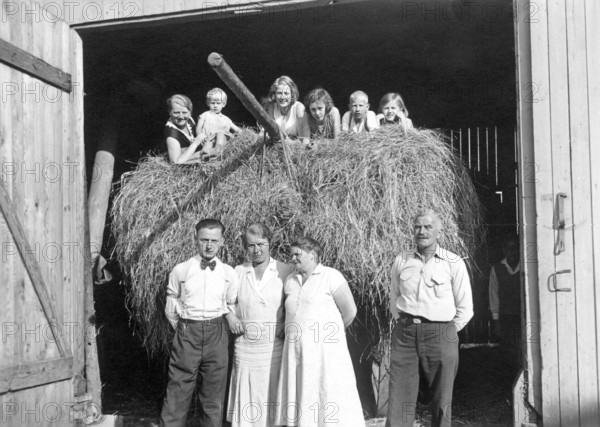 Farming family in front of the barn, ca. 1930, Family posing smiling in front of a bale of hay in