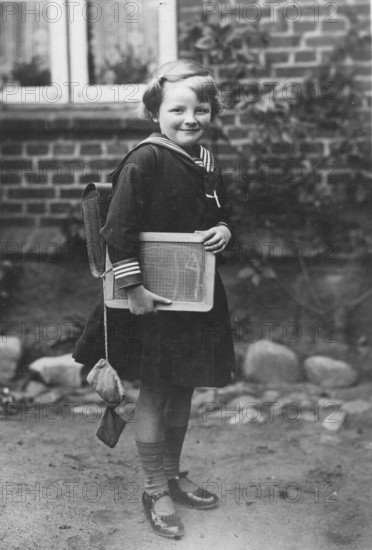 School enrolment, 1920s, A girl with a slate and school uniform stands in front of a brick wall,