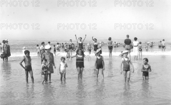 Children in the water, 1920s, Children playing in shallow water on the beach under a clear sky. A