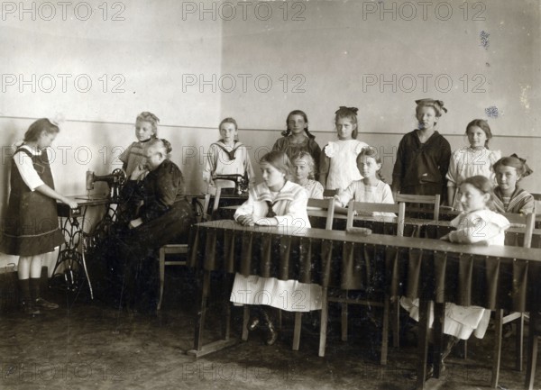 Sewing class at school, 1920, girls in a classroom practising sewing while a teacher sits at a
