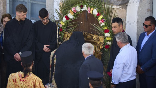 A priest prays in front of a wreath decorated with flowers, surrounded by people in traditional and
