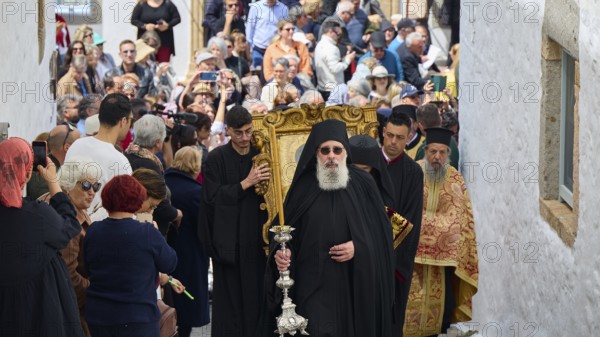 A priest leads a procession through a narrow street, followed by worshippers and surrounded by a