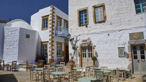 An outdoor area of a café with empty tables and chairs in front of white buildings on a sunny day,