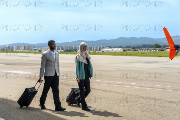 Businessman and businesswoman walking along the airfield tarmac, pulling carry on luggage while