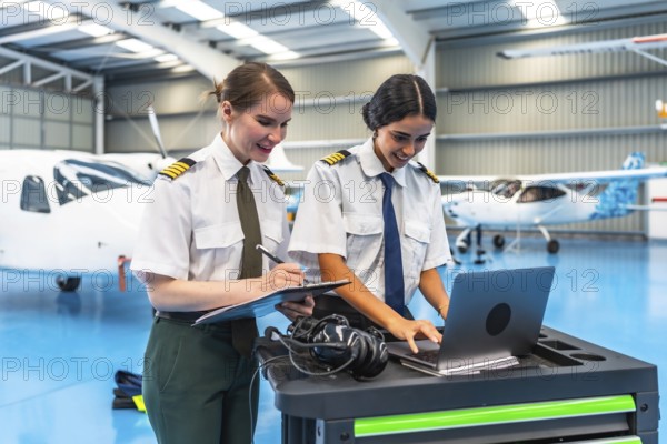 Two female pilots in uniform working on a laptop and clipboard in a spacious aircraft hangar,