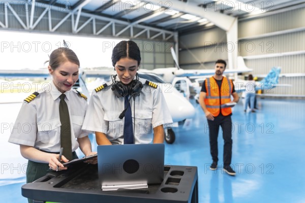 Two female colleagues pilots preparing flight plan using laptop and writing on clipboard in hangar