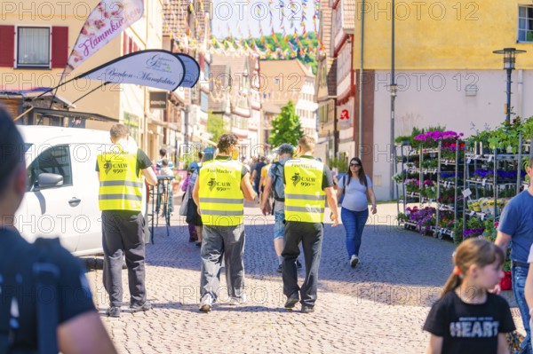 Three men in yellow waistcoats walk along a busy street, surrounded by people, Calw railway station