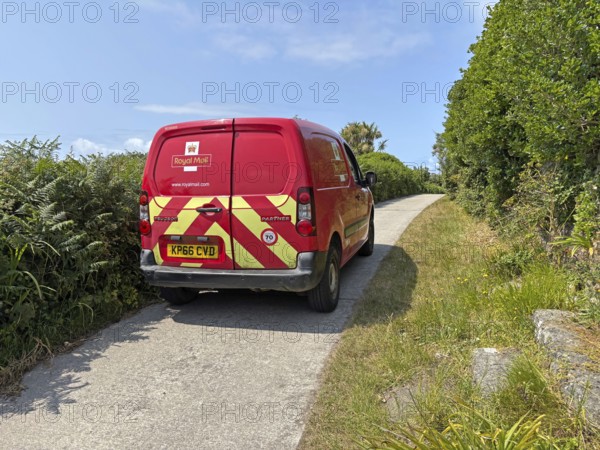 A red Royal Mail van on a rural road surrounded by lush greenery under a sunny sky, St Agnes, Isles