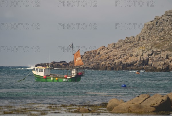 Small boat with orange sail in calm coastal waters near rocky cliff, Tresco, Isles of Scilly,