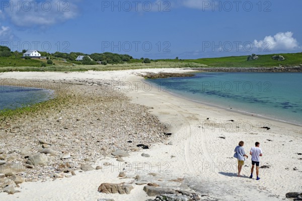 Two people walking along a soft sandy beach with blue sea and sky, St Agnes, Isles of Scilly,