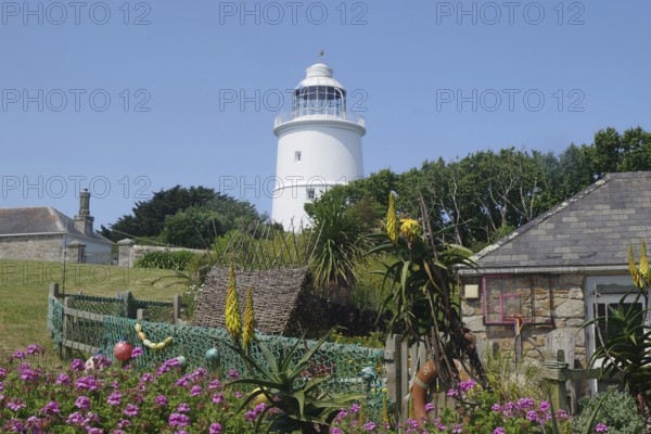 A lighthouse next to a sunny garden with blooming flowers and stone houses under a blue sky, St