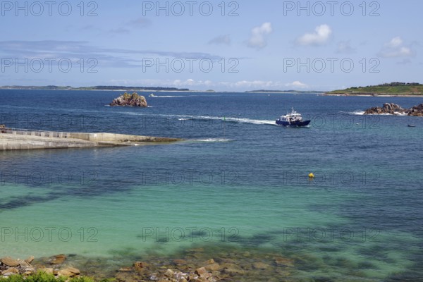 A boat sails on the open sea between small islands, under a clear blue sky, St Agnes, Isles of