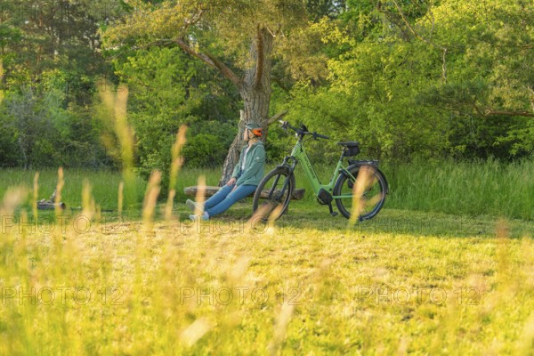 Cyclist sitting relaxed next to a bicycle under a tree on a green meadow, e-bike excursion,