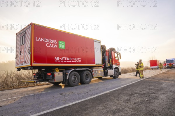 A large red fire truck is parked on a landscape road in the morning light, firefighters next to it,