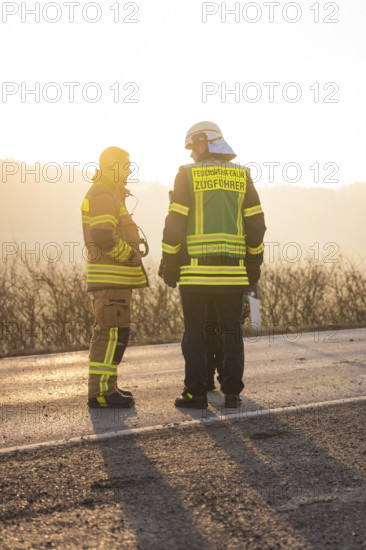 Two firefighters in protective clothing stand on a street at sunset, fire brigade exercise on the