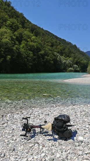 A bicycle is lying on a pebble beach by a river with clear water and wooded banks, Soca Valley,
