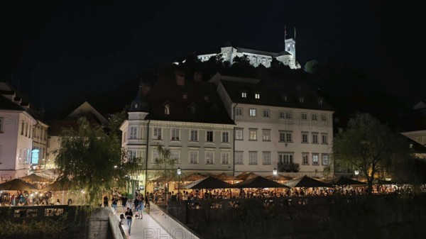 Castle illuminated at night surrounded by illuminated restaurants and visitors in an urban setting,